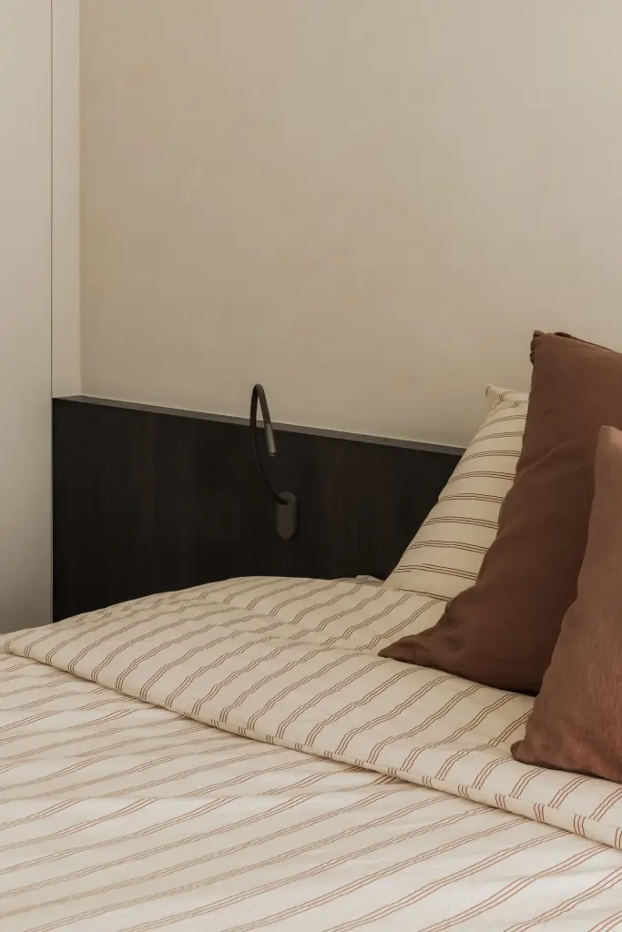 Bed met leeslamp en kussens naast een donker nachtkastje, Appartement in Nieuwpoort, interieur Laura Calleeuw