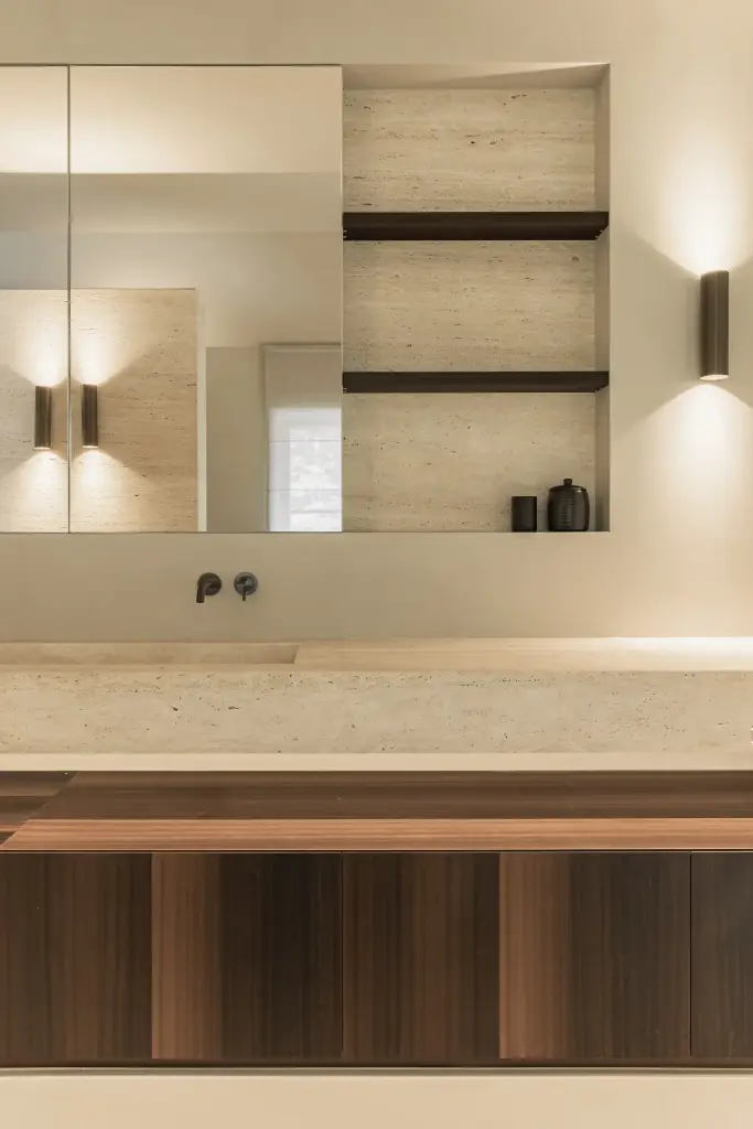 Wastafel in travertijn onder een spiegelkast, met houten onderkast en wandlamp, Gezinsvilla in Heusden-Zolder, interieur Laura Calleeuw