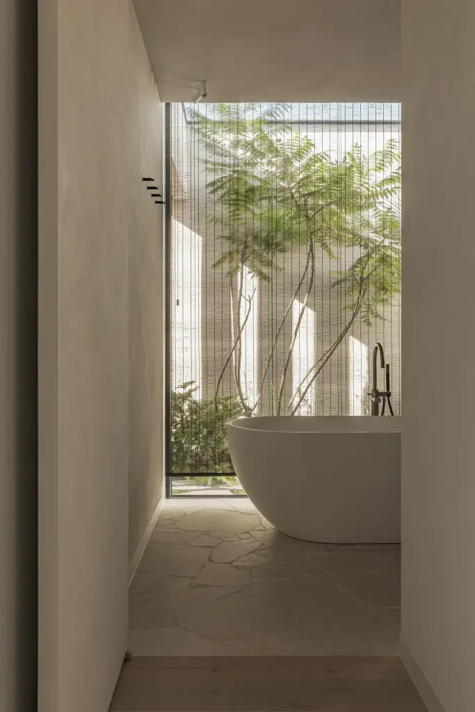 Badkamer met vrijstaand bad en zicht op een patio met boom door een hoog venster, a home that invites you to slow down, interieur Laura Calleeuw