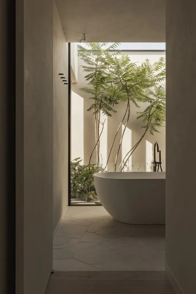 Badkamer met vrijstaand bad en zicht op een patio met boom door een hoog venster, a home that invites you to slow down, interieur Laura Calleeuw