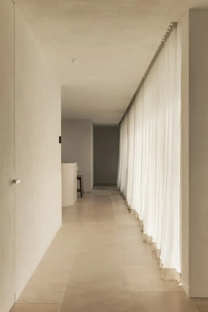 Lange gang met lichte natuurstenen vloer en vitrage langs de ramen, a home that invites you to slow down, interieur Laura Calleeuw