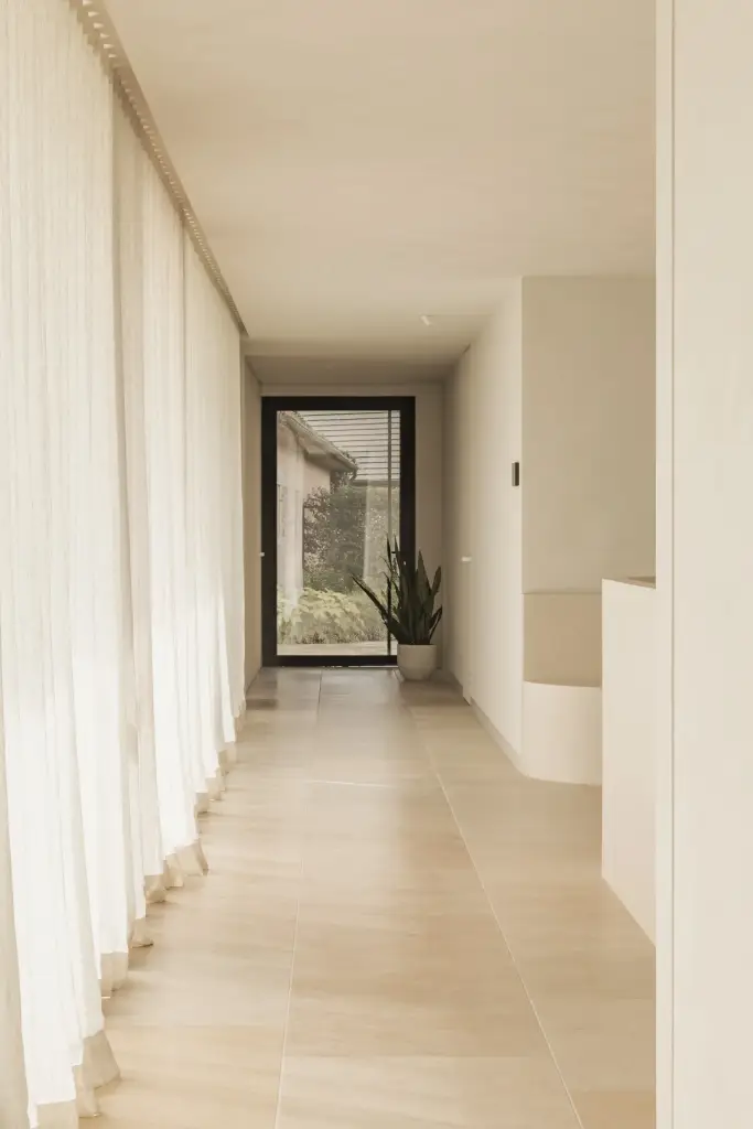 Gang met lange vitrage, lichte vloer en een glazen deur met plant op het einde, a home that invites you to slow down, interieur Laura Calleeuw