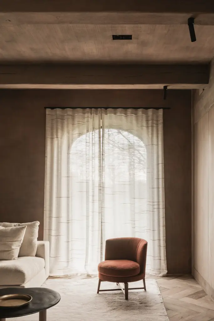 Boogvormig raam met vitrage boven een roestkleurige fauteuil en sofa, Hoeve in Westkapelle, interieur Laura Calleeuw