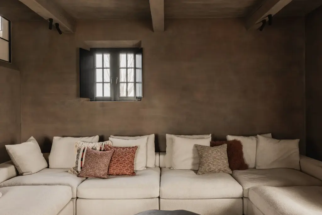 Zithoek met witte hoeksofa, kussens en een bruine leempleisterwand onder houten balken, Hoeve in Westkapelle, interieur Laura Calleeuw