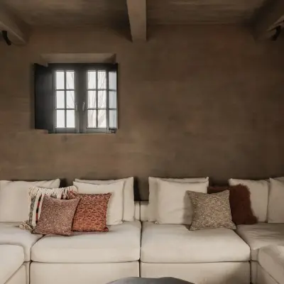Zithoek met witte hoeksofa, kussens en een bruine leempleisterwand onder houten balken, Hoeve in Westkapelle, interieur Laura Calleeuw