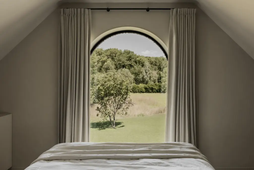 Slaapkamer met boogvormig raam, gordijnen en zicht op een boom in het landschap, a masterful balance between authenticity and comfort, interieur Laura Calleeuw