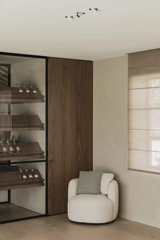 Wijnkast met flessen in staal en glas naast een witte fauteuil en roman blind, a masterful balance between authenticity and comfort, interieur Laura Calleeuw