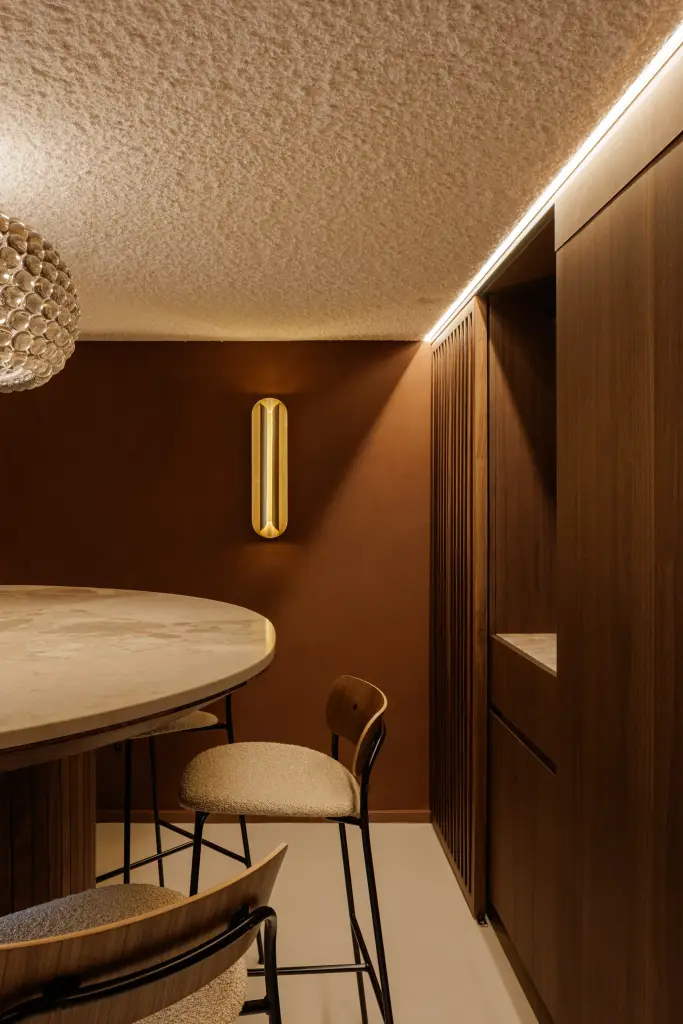 Eethoek met ronde natuurstenen tafel, barkruk en een bruine pleisterwand met gouden wandlamp, Villarenovatie in Knokke, interieur Laura Calleeuw