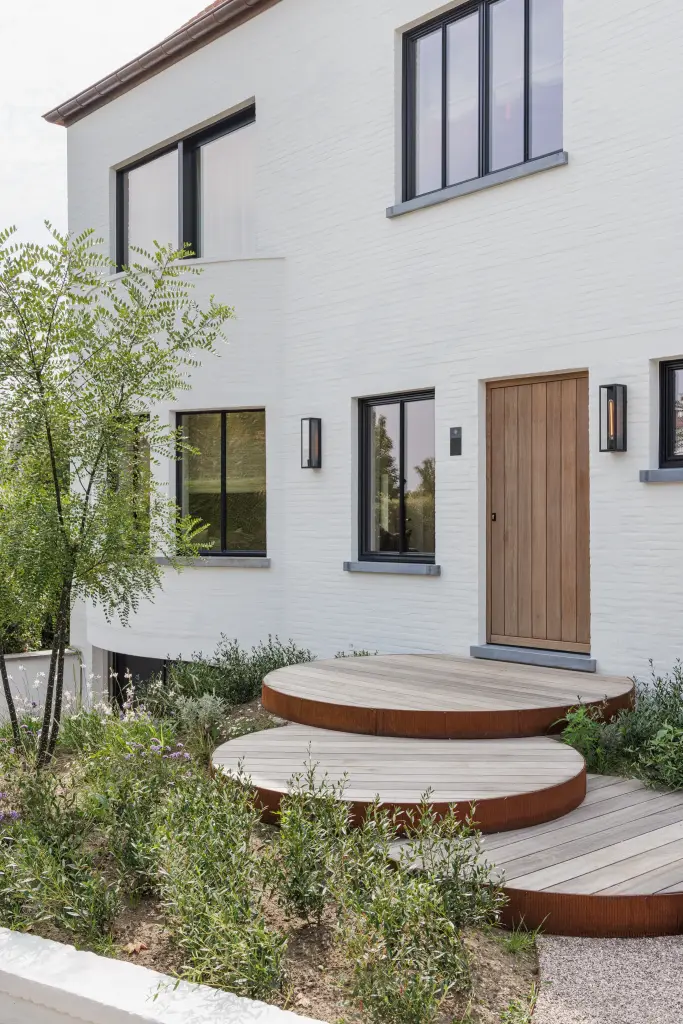Gevel van de witte woning met houten voordeur, ronde houten trappen en beplante tuin, Villarenovatie in Knokke, interieur Laura Calleeuw
