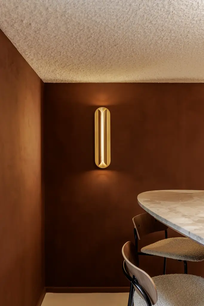 Gouden wandlamp op een bruine pleisterwand boven een halfronde natuurstenen tafel, Villarenovatie in Knokke, interieur Laura Calleeuw