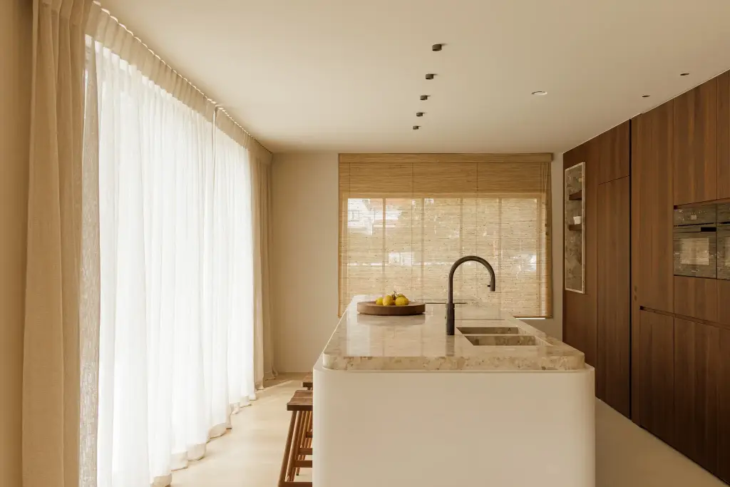 Keuken met natuurstenen kookeiland, notelaren kastenwand, roman blind en lange vitrage, Villarenovatie in Knokke, interieur Laura Calleeuw