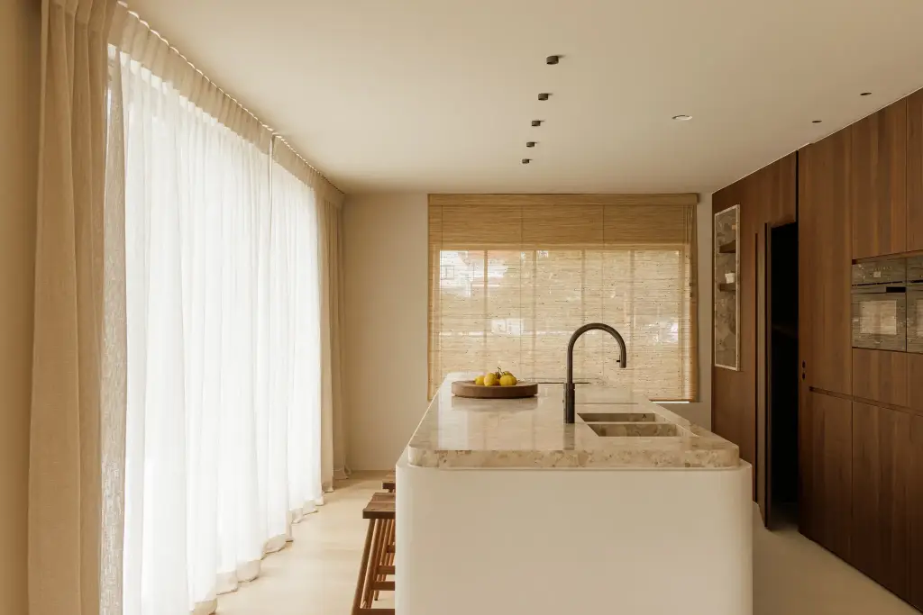 Keuken met natuurstenen kookeiland, notelaren kastenwand, roman blind en lange vitrage, Villarenovatie in Knokke, interieur Laura Calleeuw