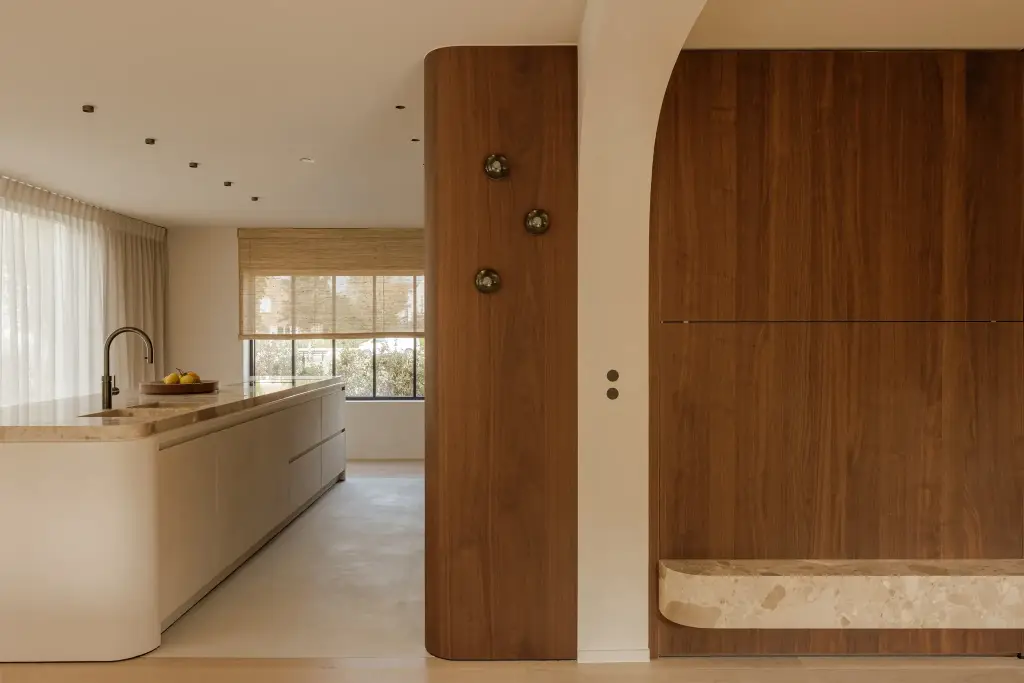 Keuken met kookeiland in lichte tint, notelaren wand met bronzen knoppen en groot raam met vitrage, Villarenovatie in Knokke, interieur Laura Calleeuw