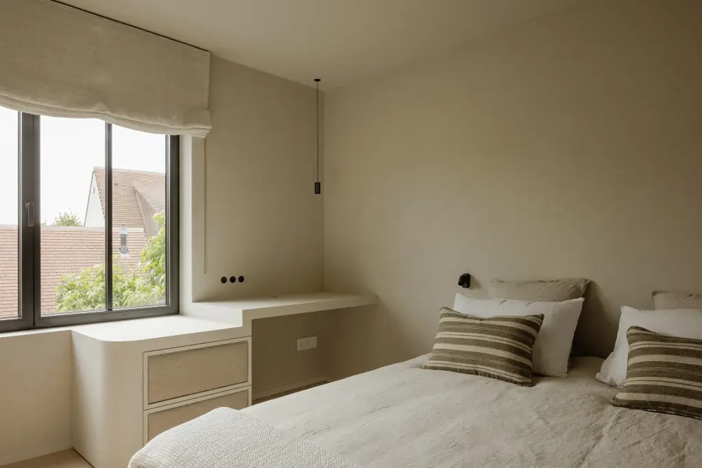 Slaapkamer met bed, ingebouwd bureau onder het raam en een roman blind, Villarenovatie in Knokke, interieur Laura Calleeuw