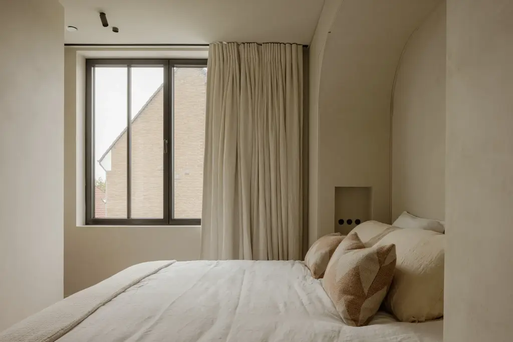 Slaapkamer met bed, kussens, zwart raam met gordijn en een boogvormige nis, Villarenovatie in Knokke, interieur Laura Calleeuw