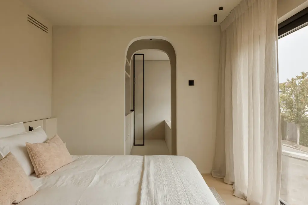 Slaapkamer met een boogvormige doorgang met stalen glasdeur naar de badkamer, bed en vitrage, Villarenovatie in Knokke, interieur Laura Calleeuw