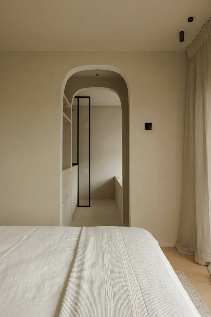 Slaapkamer met een boogvormige doorgang met stalen glasdeur naar de badkamer, bed en vitrage, Villarenovatie in Knokke, interieur Laura Calleeuw