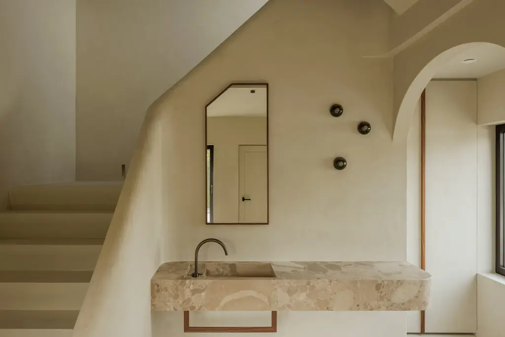 Natuurstenen wastafel met wandkraan, hoge spiegel en bronzen wandlampjes tegen kalkpleister, Villarenovatie in Knokke, interieur Laura Calleeuw