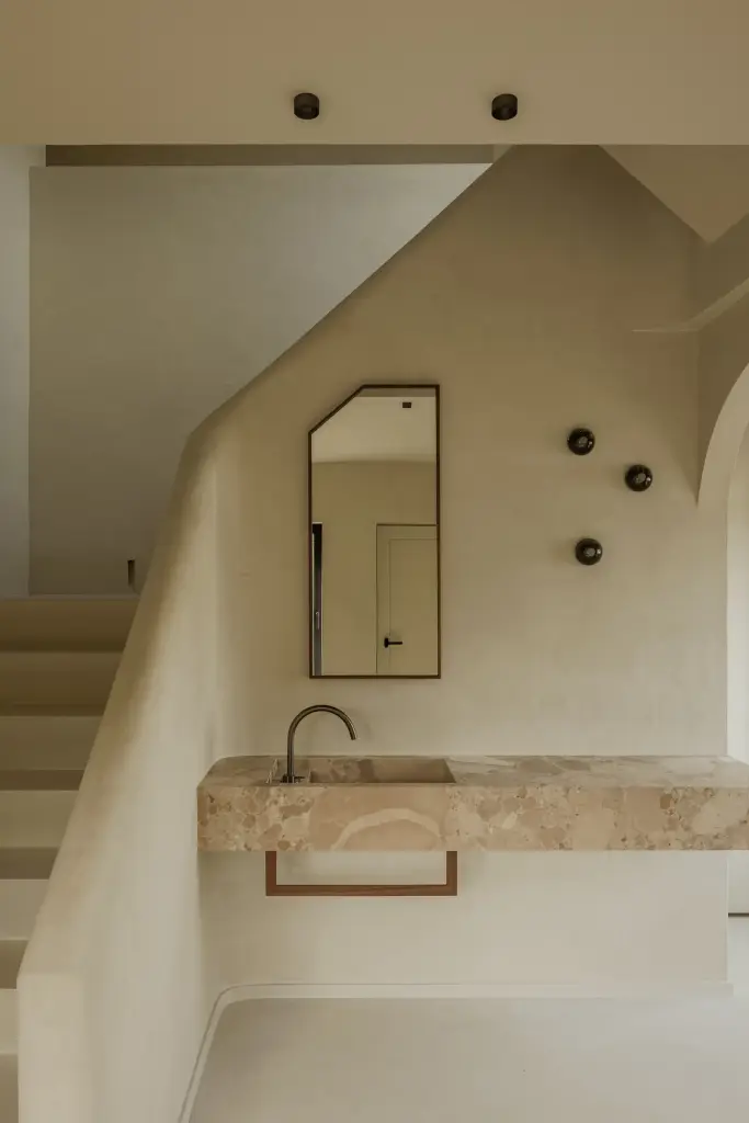 Natuurstenen wastafel met wandkraan, hoge spiegel en bronzen wandlampjes tegen kalkpleister, Villarenovatie in Knokke, interieur Laura Calleeuw