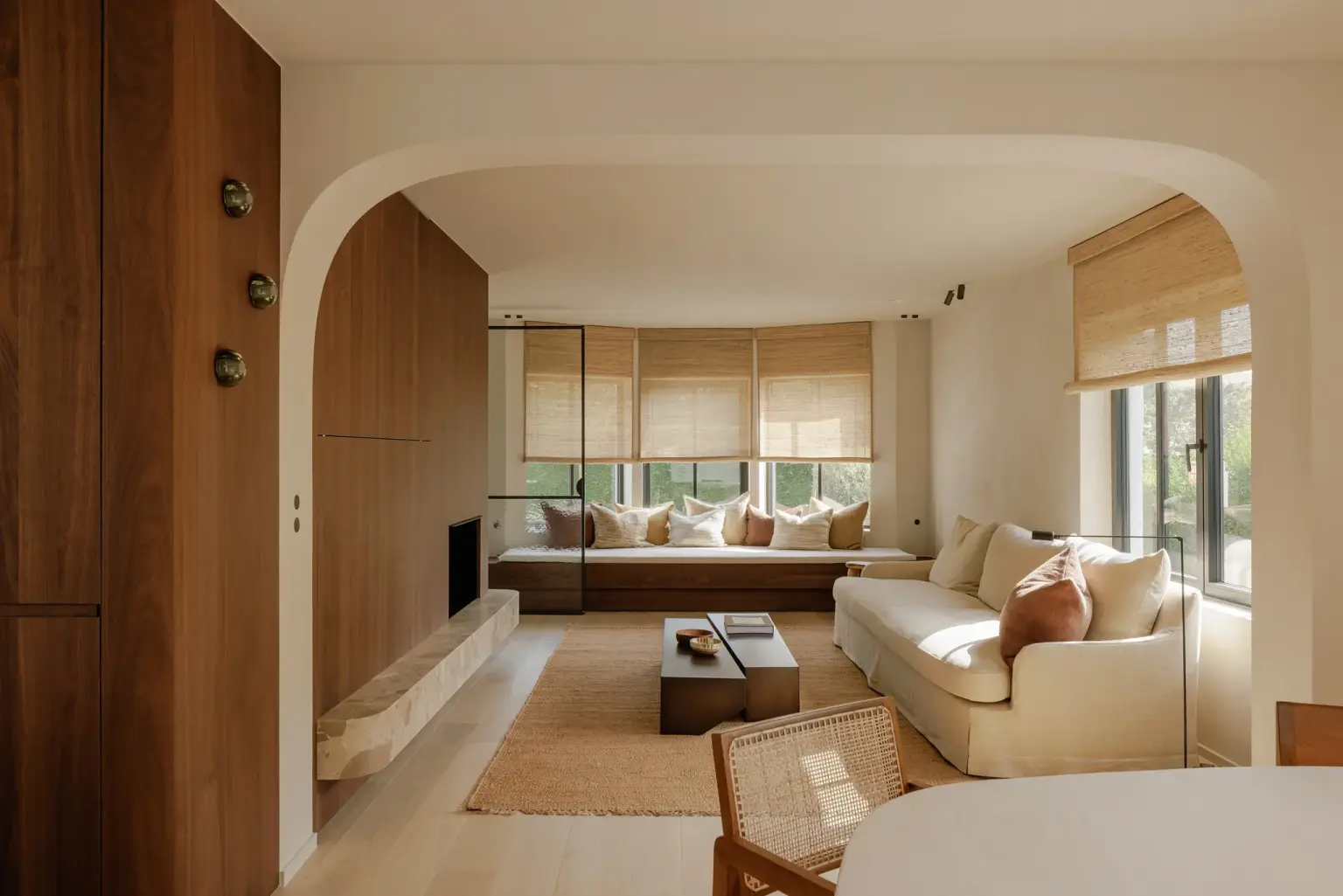 Leefruimte met boogopening, notelaren kastenwand, hoeksofa, rieten stoelen en roman blinds, Villarenovatie in Knokke, interieur Laura Calleeuw