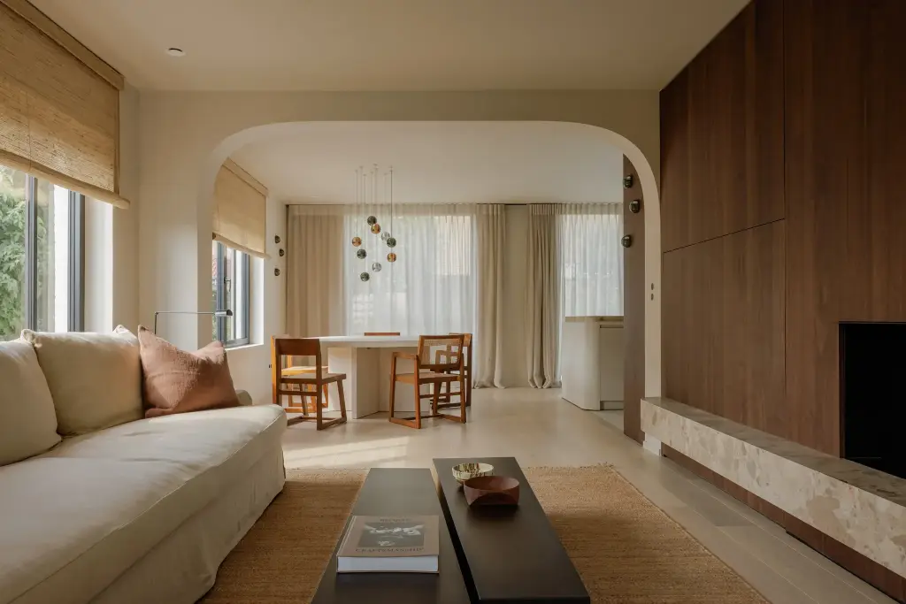 Leefruimte met lange zitbank, donkere salontafel, eethoek en een wand in notelaar met open haard, Villarenovatie in Knokke, interieur Laura Calleeuw