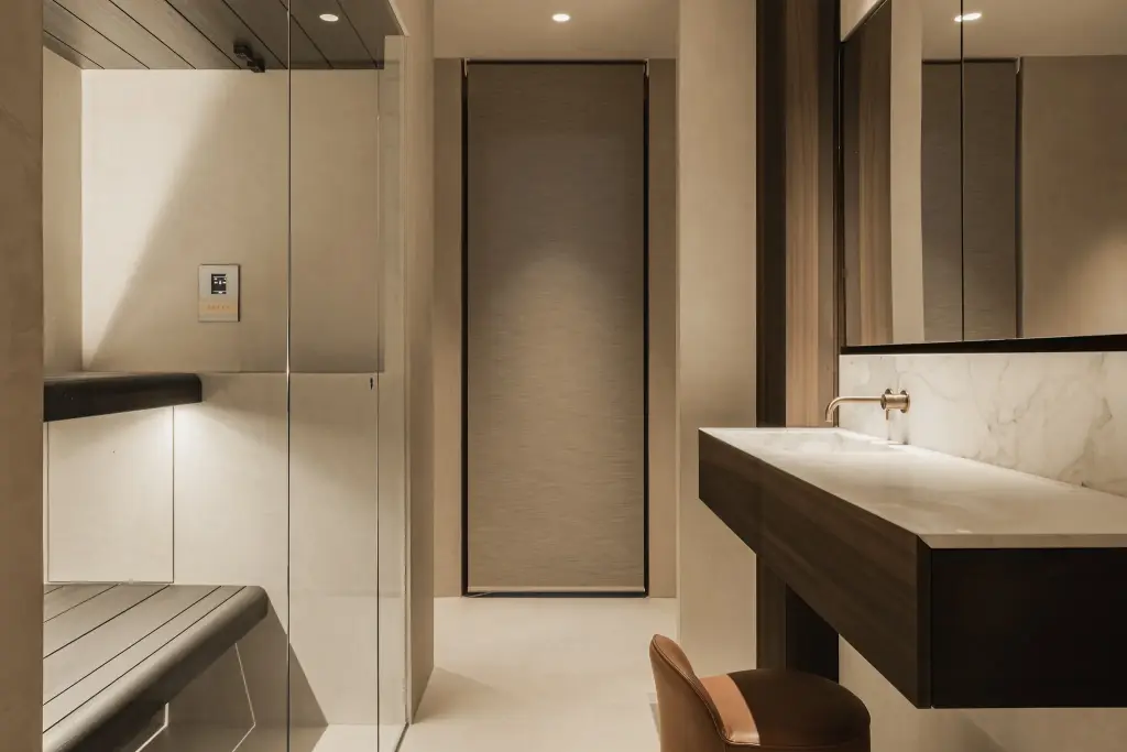 Badkamer met zwevend donker houten wastafelmeubel, marmeren wand, glazen sauna en een leren stoel, Penthouse in Kortrijk, interieur Laura Calleeuw