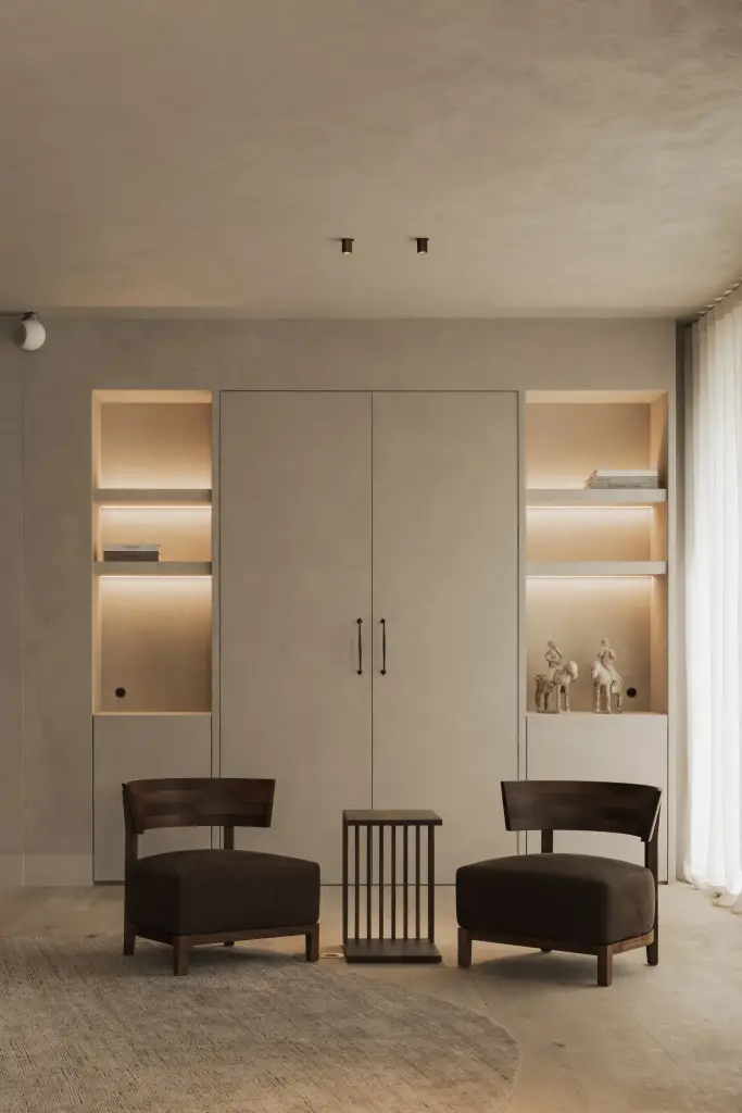 Kastenwand in kalkpleister met verlichte nissen en twee donkere fauteuils met een bijzettafeltje, Hoeve in Hansbeke, interieur Laura Calleeuw