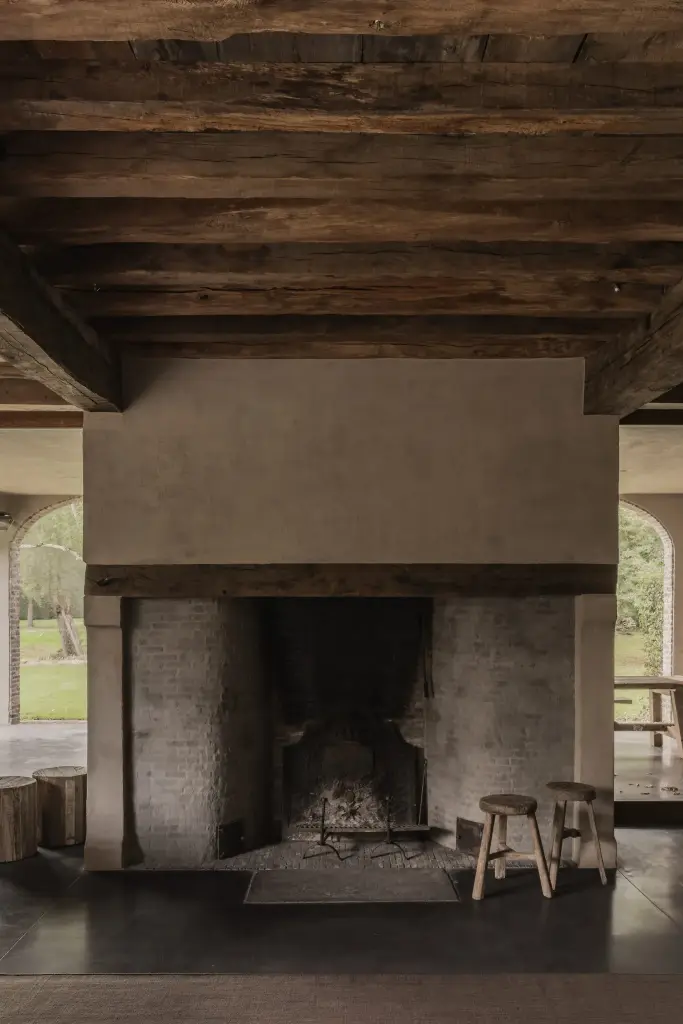 Grote open haard in kalkpleister onder oude houten balken met zicht op de tuin, Hoeve in Hansbeke, interieur Laura Calleeuw
