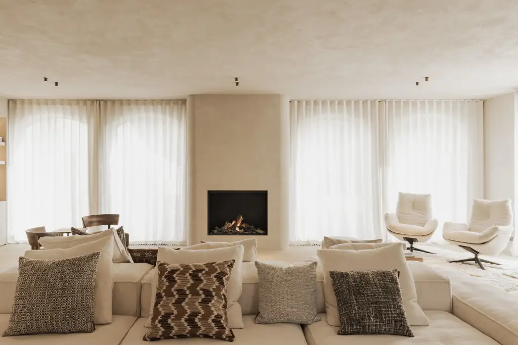 Woonkamer met hoeksofa, kussens, open haard in een gepleisterd volume, fauteuils en boogvormige vensters, Hoeve in Hansbeke, interieur Laura Calleeuw