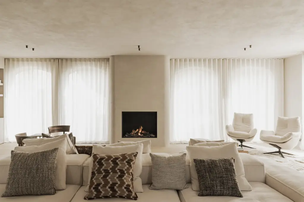 Woonkamer met hoeksofa, kussens, open haard in een gepleisterd volume, fauteuils en boogvormige vensters, Hoeve in Hansbeke, interieur Laura Calleeuw