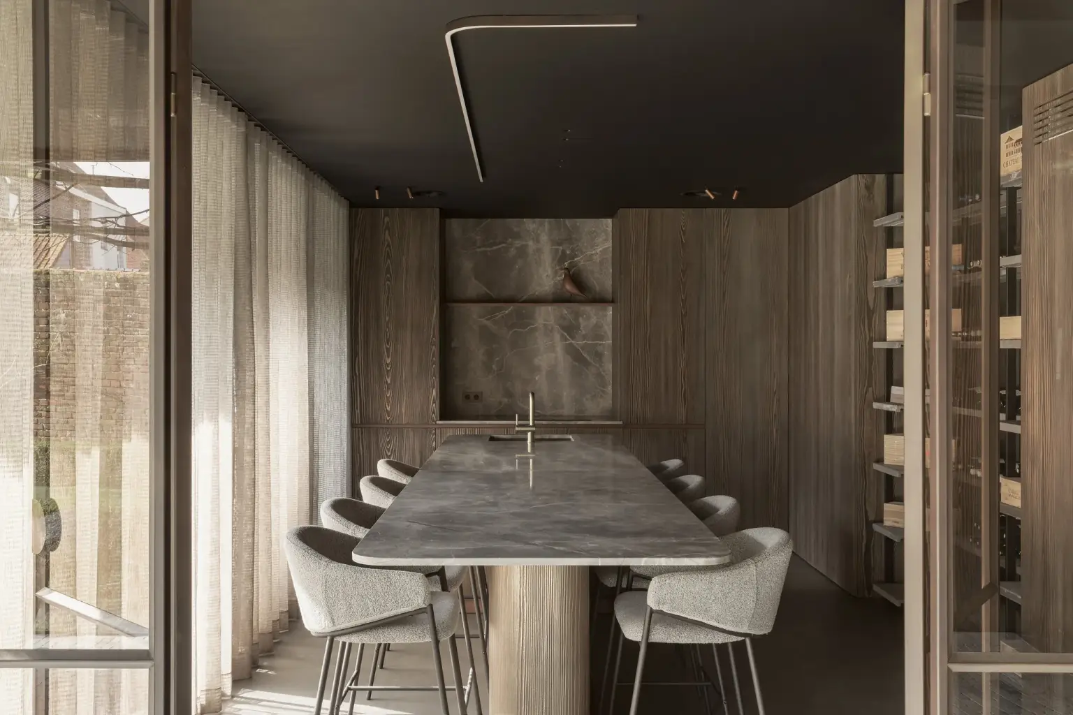 Lange natuurstenen tafel met witte stoelen in een donkere ruimte met stalen glaswanden en wijnrekken, a space that changes with the rhythm of the day, interieur Laura Calleeuw