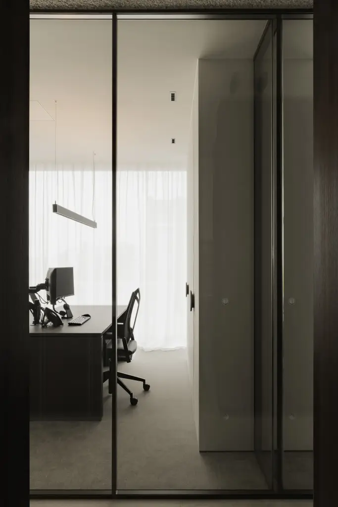 Stalen glazen wand met doorkijk naar het kantoor met bureaus en een lange lamp, Kantoor KBC in Ardooie, interieur Laura Calleeuw