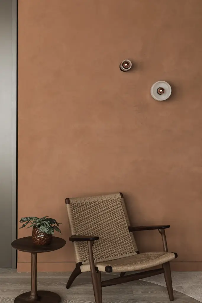 Rieten fauteuil met houten bijzettafel, plant en ronde wandlampjes tegen de terracotta wand, Kantoor KBC in Ardooie, interieur Laura Calleeuw