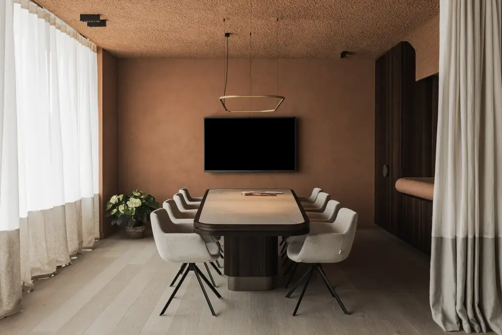 Vergaderruimte met ovale tafel, witte draaistoelen, televisie en terracotta pleisterwand achter lange gordijnen, Kantoor KBC in Ardooie, interieur Laura Calleeuw