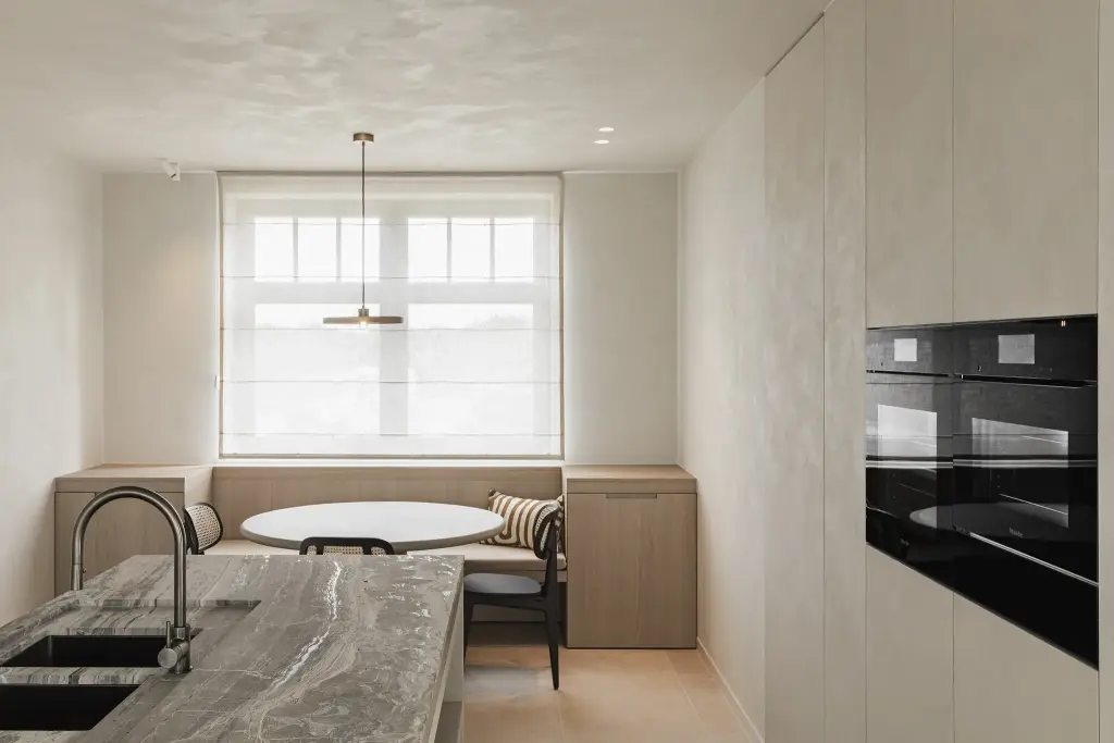 Keuken met travertijn eiland, inbouwovens en eethoek met zitbank onder het raam, where architecture creates space for experience, interieur Laura Calleeuw