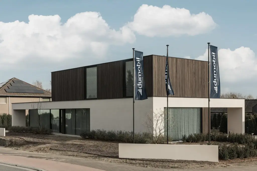 Moderne gevel van het gebouw met donkere houten lamellen en lichte crepi, met drie vlaggen aan de straatkant, Kijkwoning in Erpe-Mere, interieur Laura Calleeuw