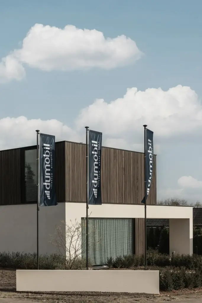 Moderne gevel van het gebouw met donkere houten lamellen en lichte crepi, met drie vlaggen aan de straatkant, Kijkwoning in Erpe-Mere, interieur Laura Calleeuw