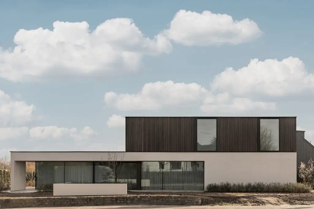 Moderne gevel van het gebouw met donkere houten lamellen en lichte crepi, met drie vlaggen aan de straatkant, Kijkwoning in Erpe-Mere, interieur Laura Calleeuw