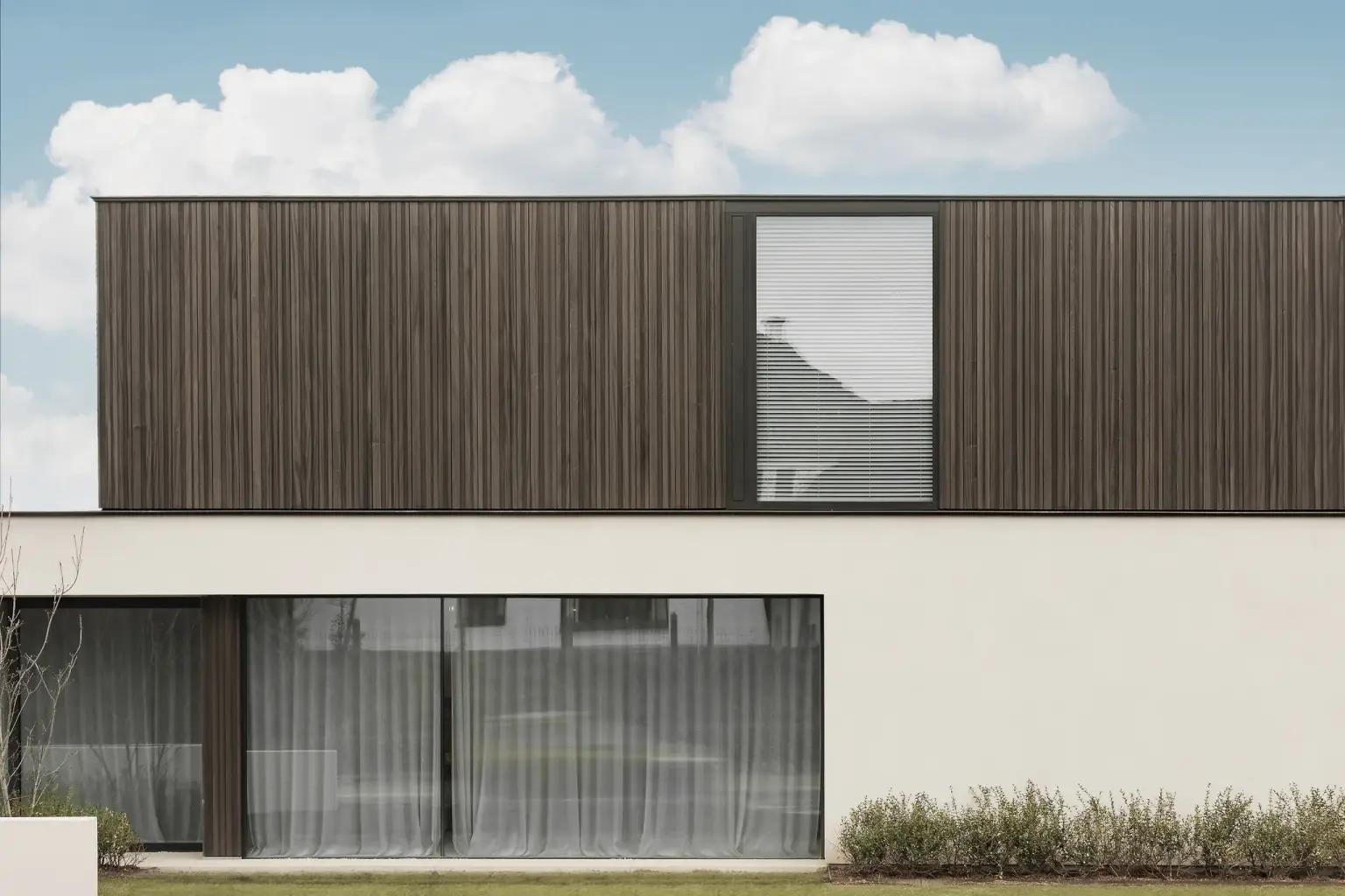 Moderne gevel van het gebouw met donkere houten lamellen en lichte crepi, met drie vlaggen aan de straatkant, Kijkwoning in Erpe-Mere, interieur Laura Calleeuw