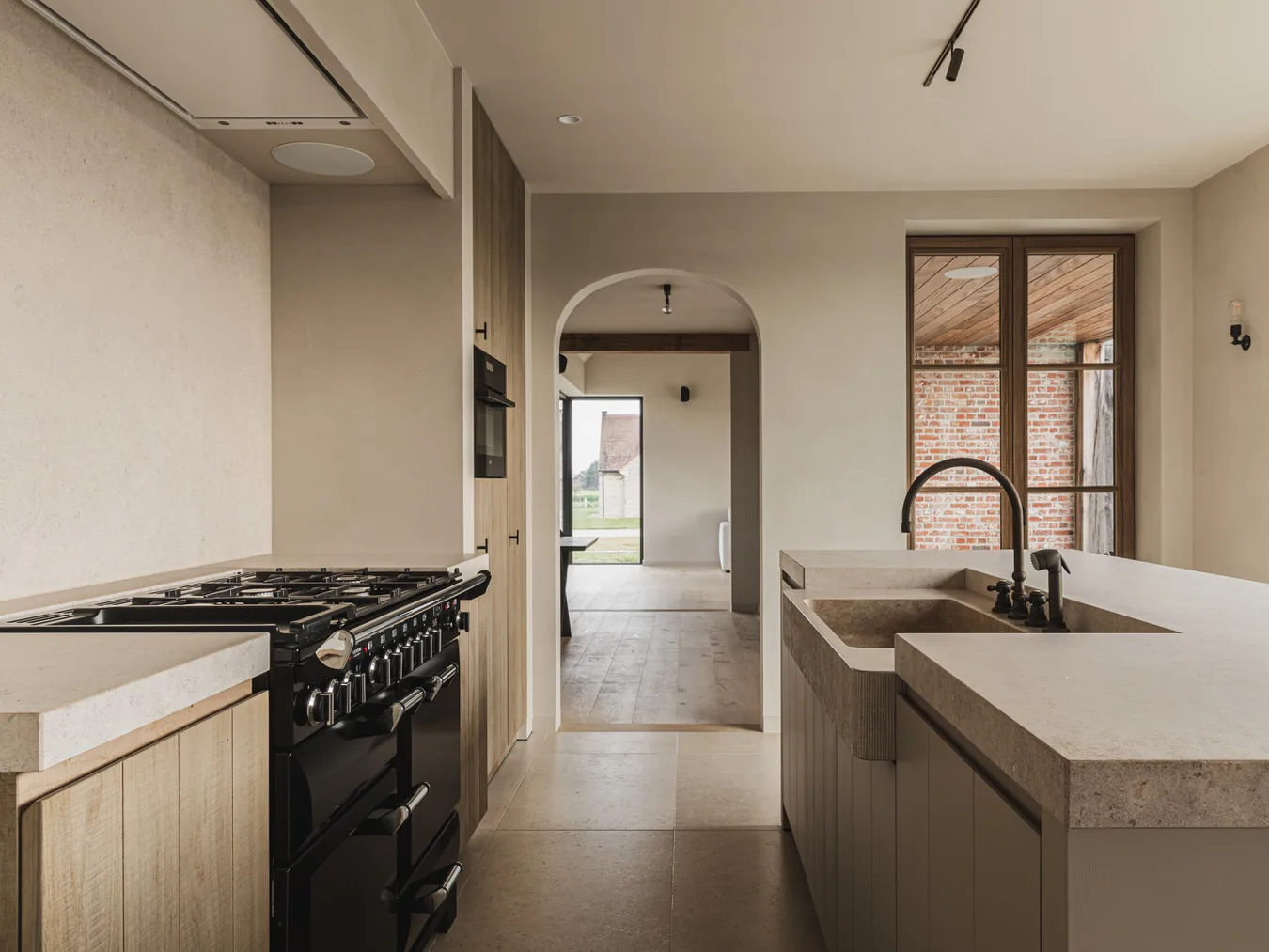 Keuken met zwart fornuis, eiland in natuursteen, doorkijk door een boogopening en eikenhouten kasten, Landelijke woning in Torhout, interieur Laura Calleeuw