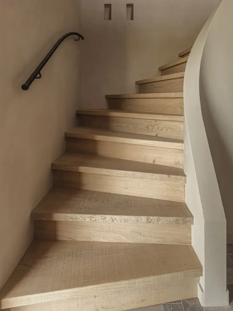 Eikenhouten trap met zwarte leuning langs een gebogen kalkpleisterwand, Landelijke woning in Torhout, interieur Laura Calleeuw