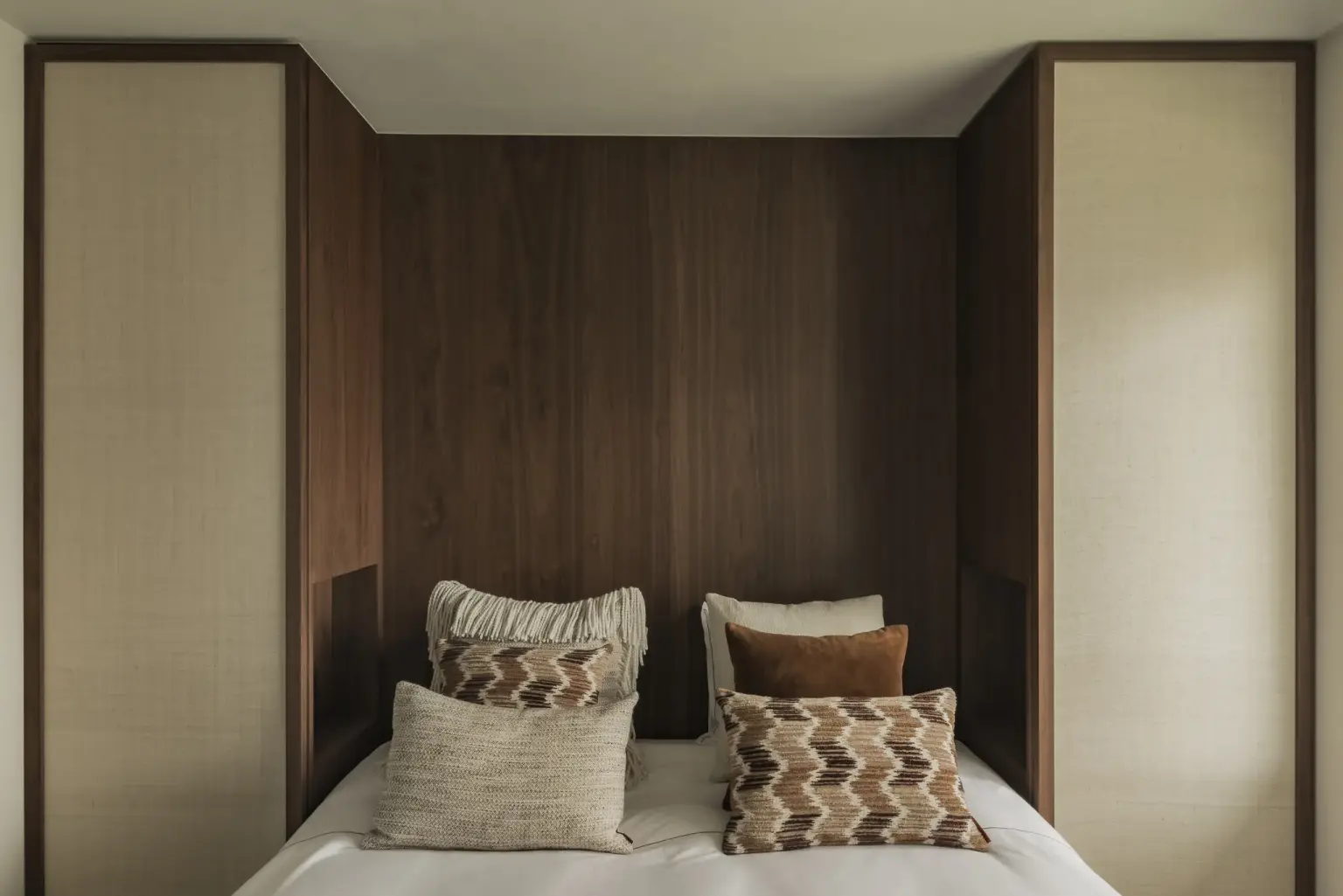 Slaapkamer met bed in walnoten nis tussen kasten met rieten panelen en sierkussens, Compact appartement in Knokke, interieur Laura Calleeuw