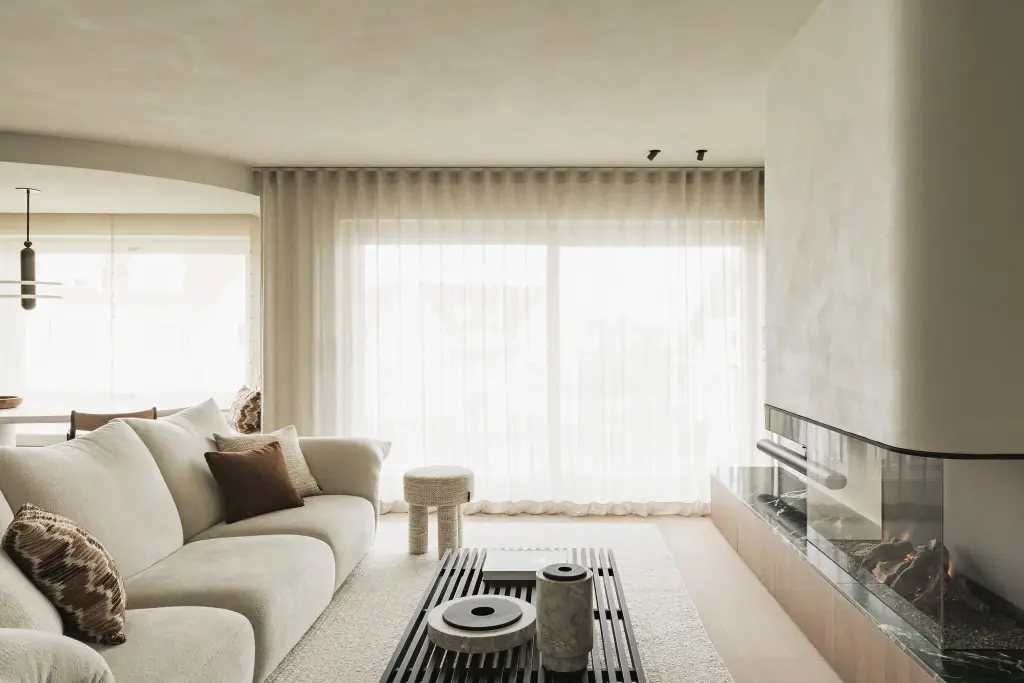 Zithoek met ronde zetel, salontafel en open haard achter linnen gordijnen, Compact appartement in Knokke, interieur Laura Calleeuw