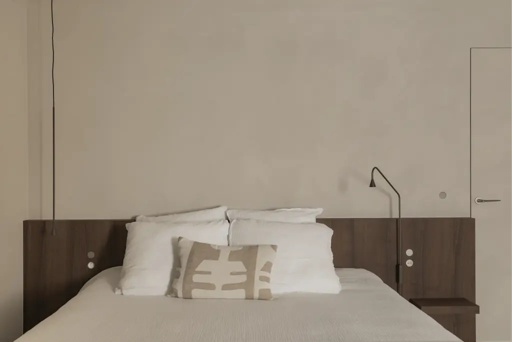 Houten hoofdbord met bed, witte kussens en een wandlamp, Het Zoute Schorre, interieur Laura Calleeuw