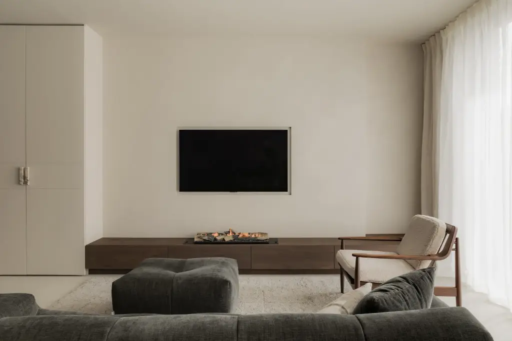 Televisie op een gepleisterde wand boven een laag houten meubel, met fauteuil en sofa, Het Zoute Schorre, interieur Laura Calleeuw
