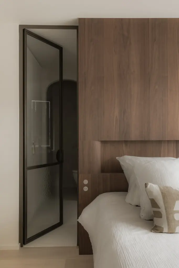 Bed met houten hoofdbord naast een stalen glasdeur naar de badkamer, Het Zoute Schorre, interieur Laura Calleeuw