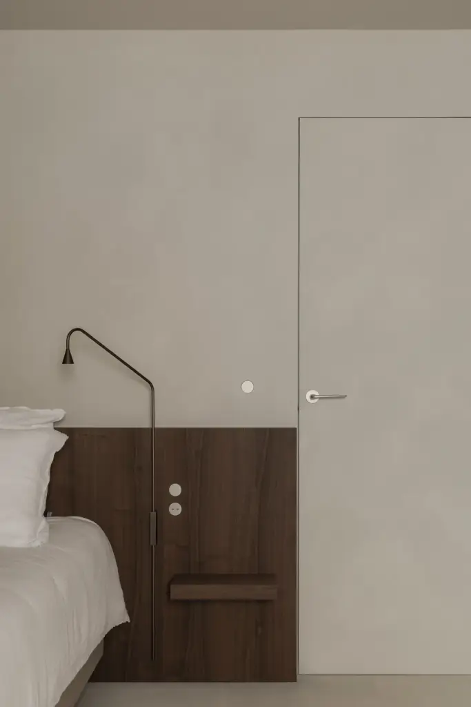 Bed met houten nachtkastje en zwarte wandlamp naast een vlakke deur, Het Zoute Schorre, interieur Laura Calleeuw