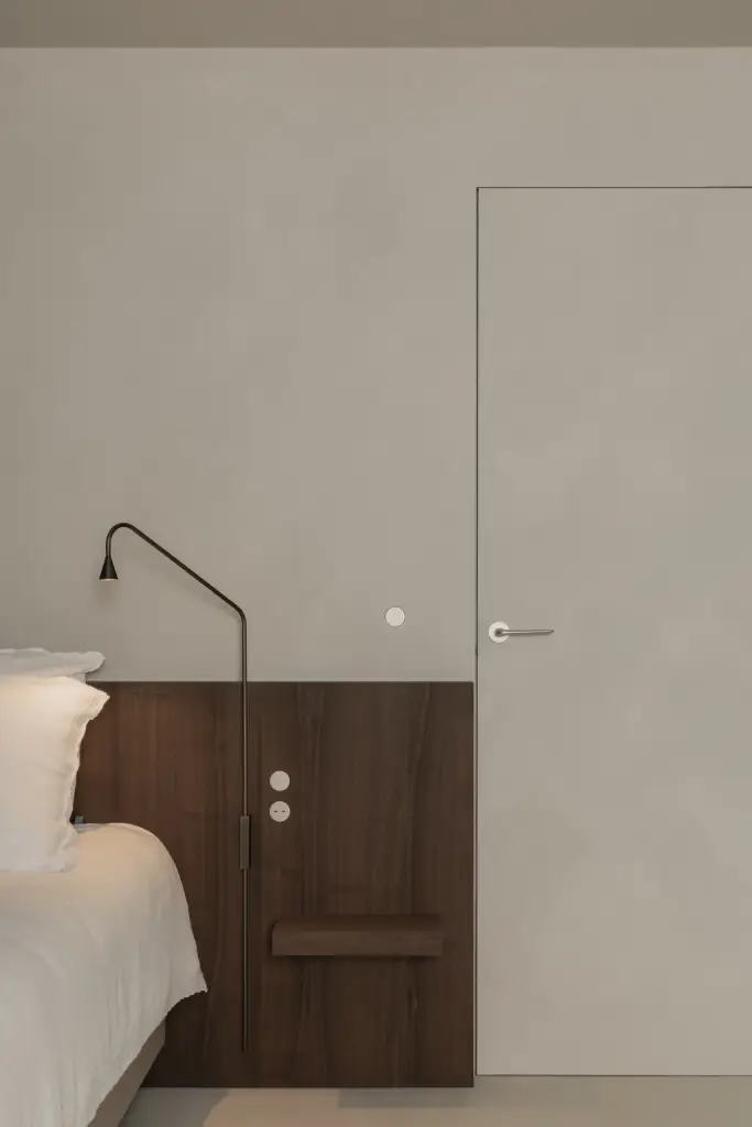 Slaapkamer met bed, houten nachtkastje, wandlamp en vlakke deur in een gepleisterde wand, Het Zoute Schorre, interieur Laura Calleeuw