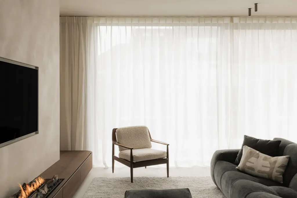 Zithoek met fauteuil, sofa en haard tegen een gepleisterde wand met televisie, Het Zoute Schorre, interieur Laura Calleeuw