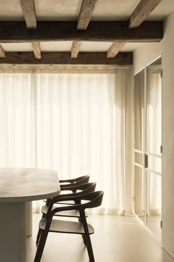 Ovale eettafel met houten stoelen voor linnen gordijnen onder een balkenplafond, Totaalrenovatie in Loppem, interieur Laura Calleeuw